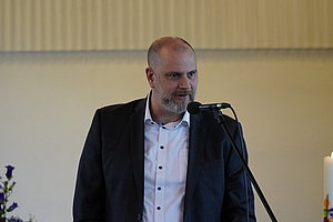 Mann in schwarzem Sakko und weißem Hemd steht vor Mikrofon in einem Raum mit Kerze und Blumen im Hintergrund.