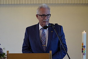 Mann im blauen Anzug mit Krawatte steht hinter einem Rednerpult mit Mikrofon, rechts eine weiße Kerze, links Blumenarrangement.