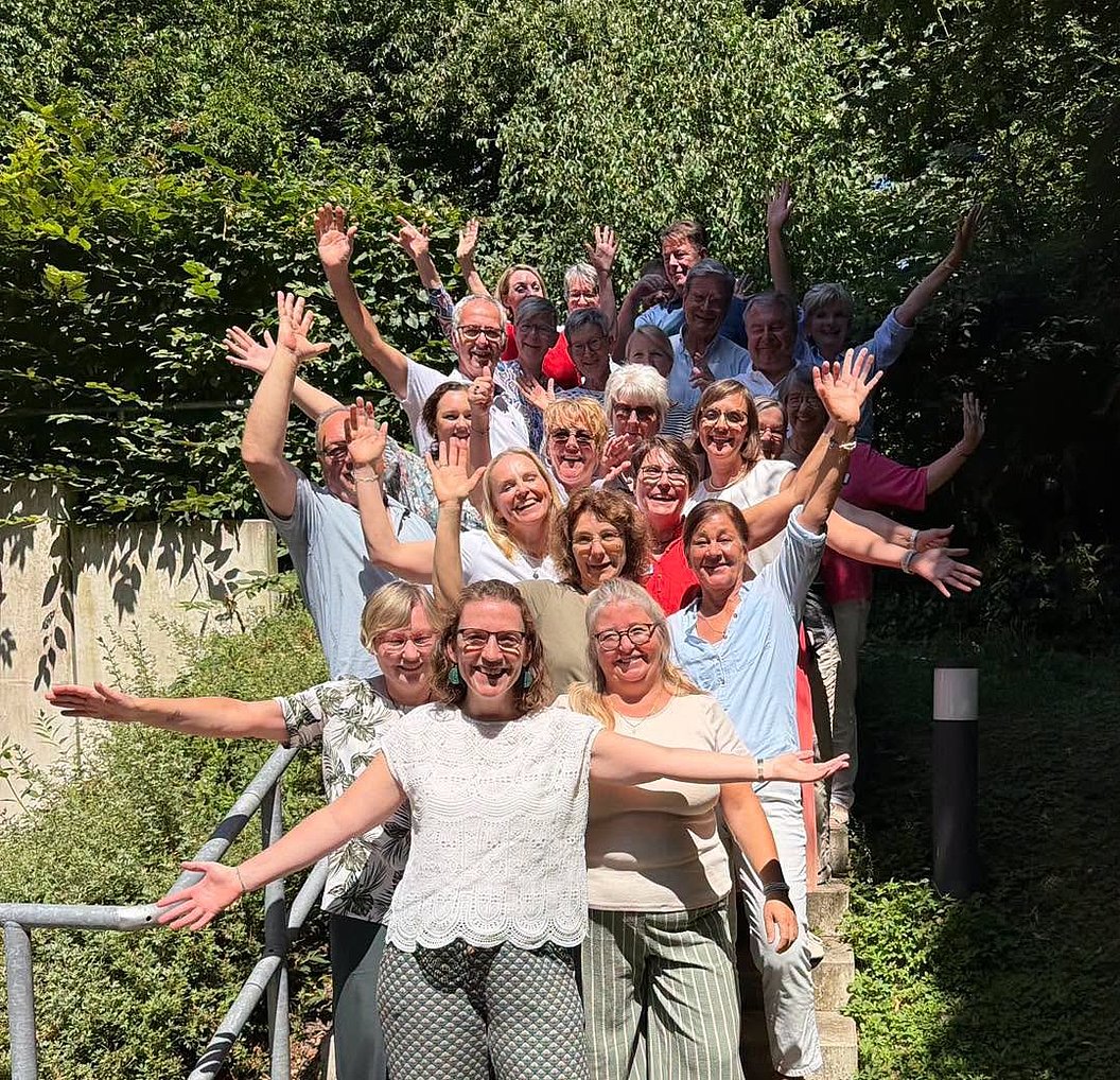 Gruppe von Menschen steht auf einer Treppe, die Arme weit geöffnet, im Freien vor grünem Gebüsch