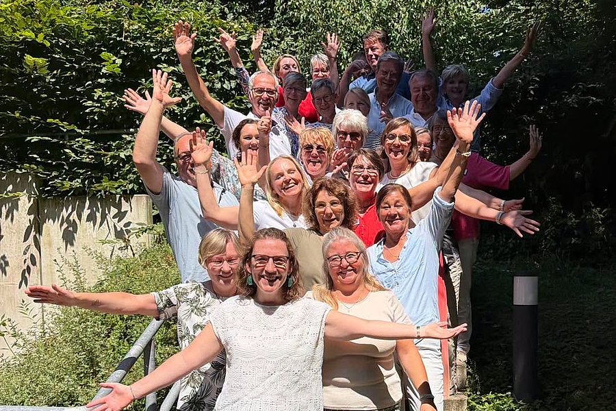 Gruppe von Menschen steht auf einer Treppe, die Arme weit geöffnet, im Freien vor grünem Gebüsch