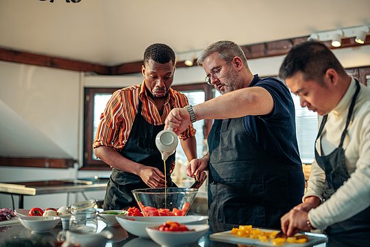 Drei Personen mit Schürzen bereiten gemeinsam in einer Küche Speisen zu, eine gießt Flüssigkeit in eine Glasschüssel mit Tomaten
