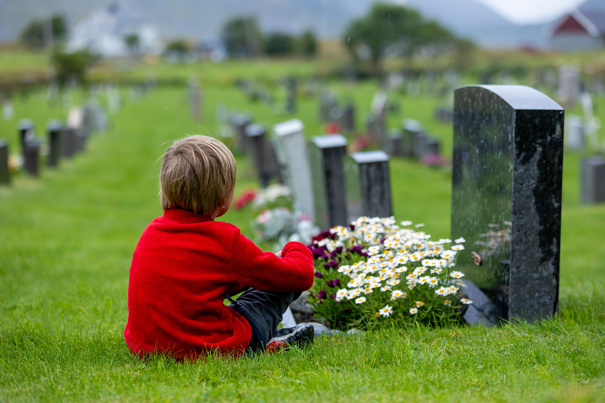 Kind mit rotem Pullover sitzt auf Gras vor einem Grabstein auf einem Friedhof